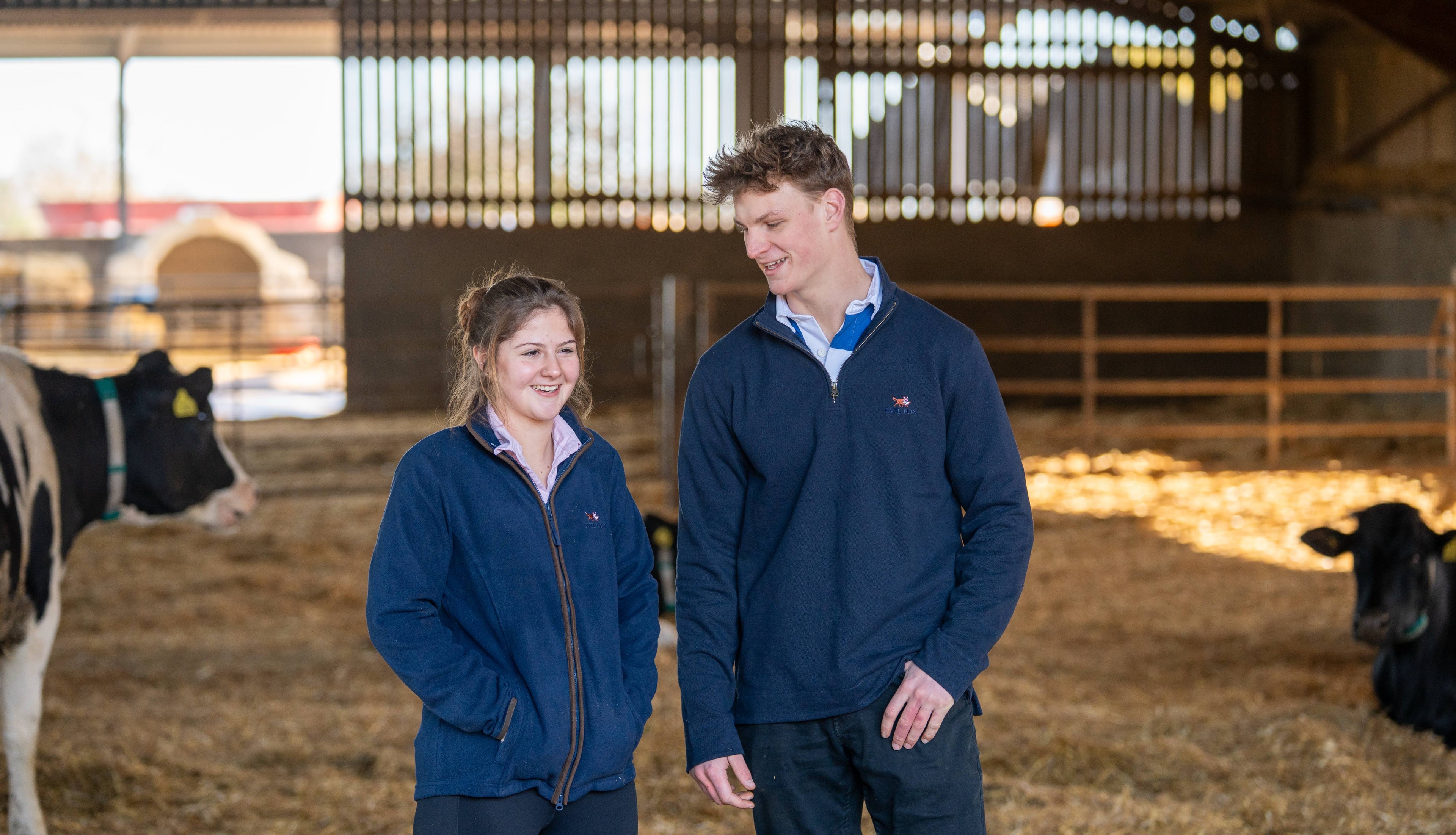Two people standing in a barn with cows in the background, wearing the evil fox quarter zip, and the evil fox female fleece.